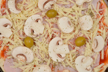 A close-up, top-down shot of a pizza being prepared with sliced mushrooms, green olives, and shredded cheese and ham on top of the dough