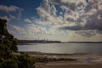 A scenic coastal view of Auckland, New Zealand, featuring the city skyline with the Sky Tower in the background, calm waters, a sandy shore, and a dramatic cloudy sky creating a moody atmosphere