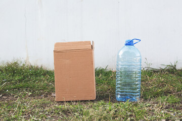 A cardboard box and a plastic water bottle placed on the grass against a white wall, representing packaging and hydration concepts