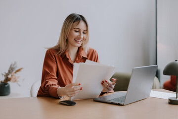 Woman sitting at desk reviews documents while smiling in a modern office setting during daytime