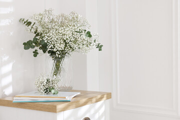 Beautiful gypsophila flowers with eucalyptus branches in vase and magazines on table at home. Space...