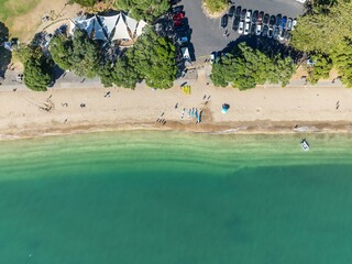 Aerial view of people enjoying a sunny day at a beach in Auckland, New Zealand. Some are swimming, sunbathing, and kayaking. A boat is anchored offshore. MISSION BAY, AUCKLAND, NZ