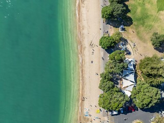 Aerial view of people enjoying a sunny day at a beach in Auckland, New Zealand. People are swimming, sunbathing, and relaxing on the sand. Trees and a park are nearby. MISSION BAY, AUCKLAND, NZ