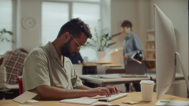 Young middle eastern man typing on computer and writing notes at desk in modern office, working on project with colleagues collaborating in background. Time lapse