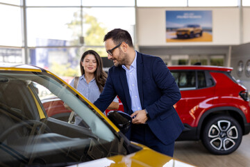 Beautiful young woman is talking to handsome car dealership worker while choosing a car in dealership, two consultants or managers in elegant suits with laptop and tablet in arms in car dealership