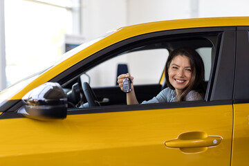 Happy brunette woman buying new cute car, raising hand up with automatic key and smiling, showroom...