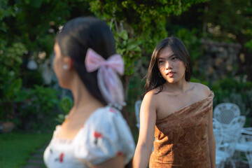 Naklejka premium Southeast Asian women in their early 20s walk hand in hand outdoors, one leading the other with a caring expression in a lush, sunlit garden