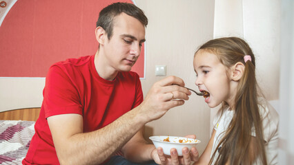 Cheerful small girl daughter and her handsome young dad spending time together at home. Father feeding his little daughter.