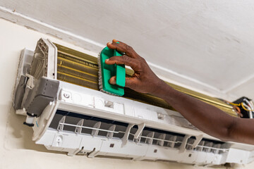 Servicing, African technician cleaning and maintaining wall-mounted air conditioning fins at home.