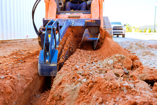 Excavation trencher equipment digs trench in red soil at construction site, highlighting industrial activity.