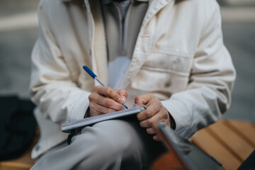 A person dressed in a light-colored jacket is sitting outside and writing in a notebook using a...
