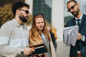 Three individuals engaged in a business discussion, analyzing documents in a collaborative setting. They appear to communicate ideas and share details while standing outdoors.
