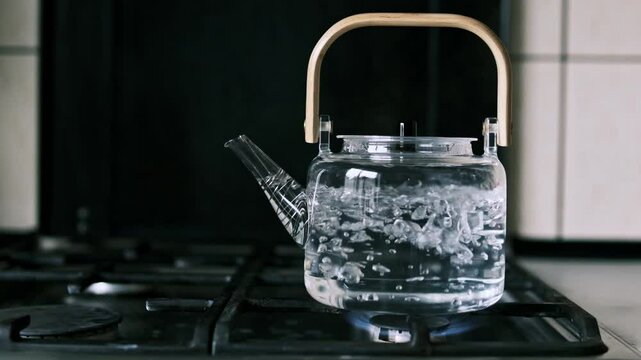 Glass kettle with a wooden handle boiling water on a gas stove in slow motion, with visible bubbles showing heat and steam. The transparent glass emphasizes the boiling process and kitchen atmosphere.