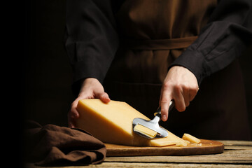 Woman cutting delicious cheese with slicer at wooden table against black background, closeup