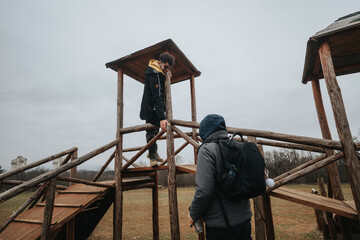 Two friends outdoors in a field exploring a wooden structure on an overcast day. The image captures...