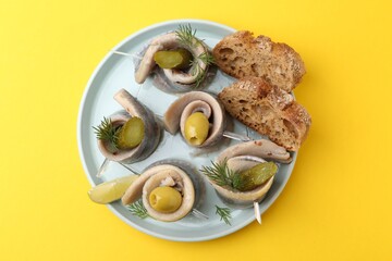 Delicious herring rolls with olives, pickles, dill and bread pieces on yellow table, top view