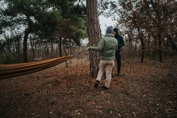 Two individuals collaborate to secure a hammock between trees in a serene forest.