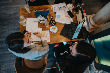 Two focused professionals working together with a laptop and documents in a cafe environment. The setting suggests teamwork, productivity, and professional conversations in a relaxed atmosphere.