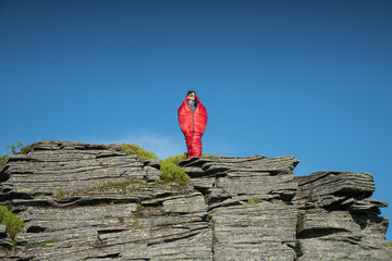 Young man wearing sunglasses and outdoor gear, standing in a red sleeping bag on a rocky mountain...