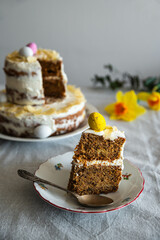 Piece of homemade two layered Easter carrot cake with ricotta cream, decorated with chocolate eggs and almonds on ornate plate and daffodils on linen tablecloth.