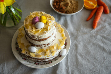 Homemade two layered Easter carrot cake with ricotta cream, decorated with chocolate eggs and almonds and carrots, oranges and almonds on linen tablecloth.