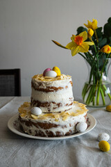 Homemade two layered Easter carrot cake with ricotta cream, decorated with chocolate eggs and almonds and daffodils on linen tablecloth.