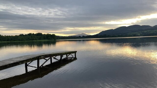 A tranquil sunset over Lake of Menteith in Scotland. A wooden dock stretches out into the calm water, reflecting the colorful sky and distant Ben A'an mountain - UK