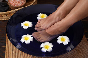 Woman soaking her feet in bowl with water and plumeria flowers on floor, closeup. Spa treatment