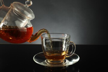 Pouring freshly brewed tea from teapot into glass cup on black table, closeup