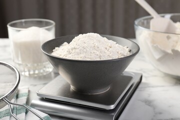 Digital kitchen scale with bowl of flour and other products on white marble table, closeup