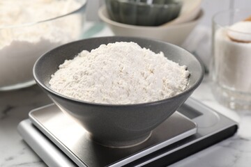 Digital kitchen scale with bowl of flour and other products on white marble table, closeup