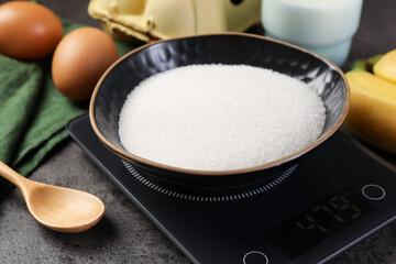 Digital kitchen scale with bowl of sugar and other products on grey table, closeup