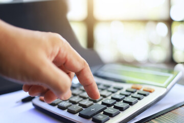 A person's hand is using a calculator on a desk to calculate income, expenses and taxes for...