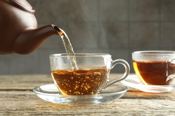 Pouring aromatic tea from teapot into cup at wooden table against grey tiled wall, closeup
