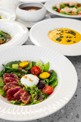 A vibrant gourmet salad featuring fresh greens, sliced tuna, cherry tomatoes, and a poached egg, elegantly presented on a white plate, complemented by a gourmet soup in the background