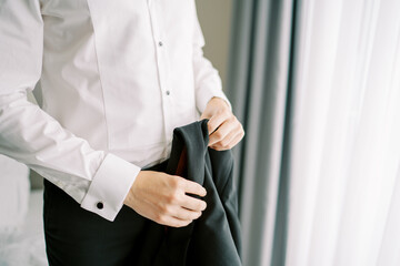 Groom holds a black jacket in his hands while standing in a hotel room. Cropped. Faceless