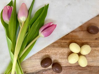 Pink tulips and white and milk chocolate eggs on a wooden and marble surface. A delicate spring-themed composition perfect for Easter, Mother's Day, or seasonal celebrations.
