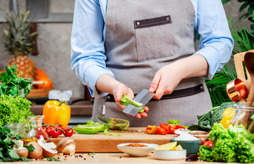 Cook cleans green paprika from seeds and membranes. Cozy kitchen with wooden table, kitchenware, vegetables, herbs and ingredients for cooking. Healthy vegan eating, culinary, recipes, food blogging