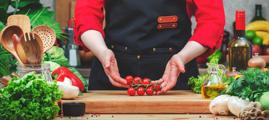 Chef holds red cherry tomatoes bunch in hands.. Cozy kitchen with wooden table, kitchenware, vegetables, herbs and ingredients for cooking. Healthy vegan eating, culinary, recipes, food blogging