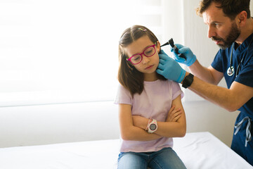 Male doctor checking ear of young female patient using otoscope during medical exam in clinic...