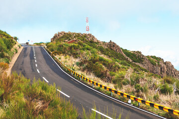 Winding road with striped guardrails curves through lush greenery, leading to atop a hill. Steep...