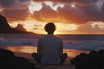 A man sits on the beach at sunset, facing away from the camera with his back to the viewer and his hands spread out in front of him
