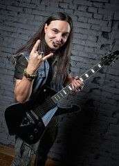 Young rock musician (headbanger) with electric guitar. Portrait of handsome long-haired man standing at wall and showing 