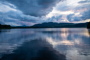 Reflection of a cloudy sky on a calm lake at dusk, surrounded by rolling hills, exuding peace and serenity - Lake of Menteith, Scotland, UK