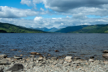 A scenic capture of Loch Katrine surrounded by majestic mountains and vivid greenery under a partly cloudy sky, located in the renowned Trossachs area of Scotland - UK