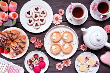Mother's Day tea table scene over a dark wood background. Assortment of sweet desserts and pastries. Above view. Pink flower and hearts theme.