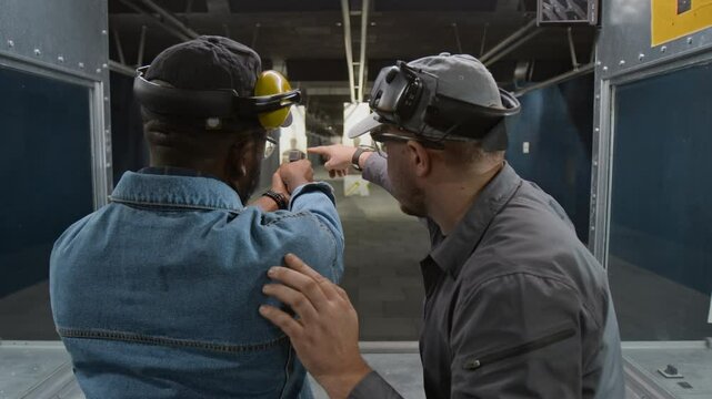 Medium rear of Caucasian male shooting coach teaching handgun aiming technique to African American male customer at indoor range, pointing to target and gunsights on weapon barrel