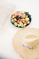 Plate of chopped fruit stands on a white sheet on the bed next to a straw hat