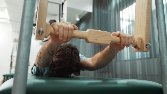 Man gripping pilates bar on reformer for upper body strength and control training during a workout at gym. Trainer doing swan dive on the cadillac for improving posture and core stability. Habituate.