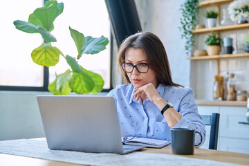 Middle-aged serious woman working at computer laptop in home office